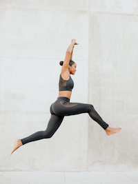 a Black model wearing matching black sports bra and yoga pants and jumping in the air